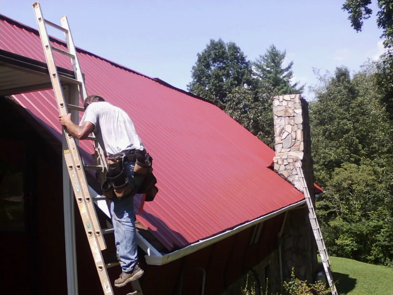 Roofer on a ladder beside a red metal roof and stone chimney — Soffit & Fascia Repair by Roofing Brothers in Glendora
